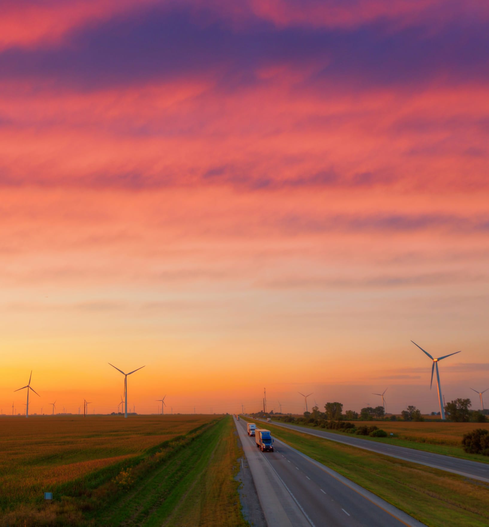 Semi trucks driving under a beautiful painted sky with wind turbines in the background.
